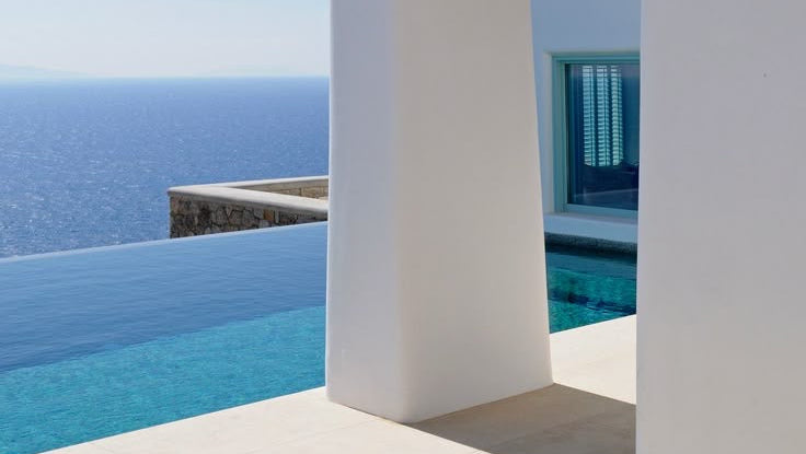 Modern white Greek architectural structure overlooking a blue infinity pool and the Aegean ocean, with a clear blue sky in the background.